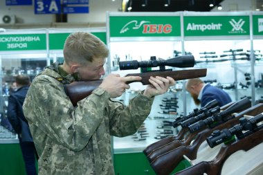 Askeri üniformalı genç bir adam teleskopik olarak bir tüfek ve silah dükkanına bakıyor. 08 Ekim 2019. Kiev, Ukrayna