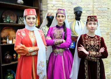 Geleneksel Tatar elbiseli üç genç kadın ayakta duruyor, arka planda mutfak aletleri var.