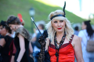 30 'lu yaşlarda, eski moda elbise ve saç kesimi giymiş orta yaşlı bir kadın elinde uzun bir ağızlık ile sokakta dikiliyor.