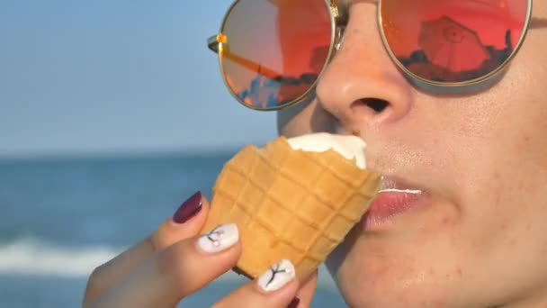 Молодая брюнетка лижет мороженое на пляже, синее море фон и солнцезащитные очки . — стоковое видео