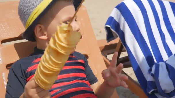 Niño comiendo papas fritas caseras en un palo, en la playa del mar. Se pone en una tumbona . — Vídeos de Stock