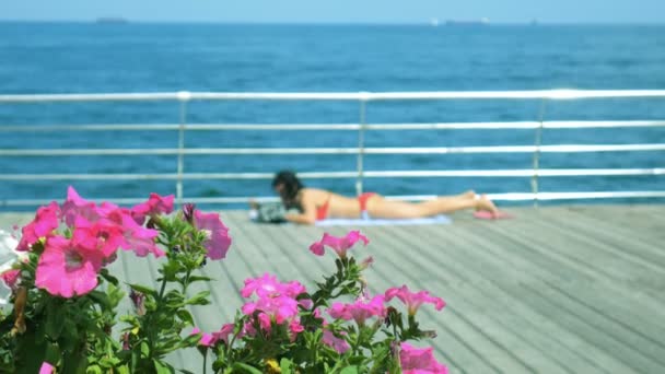 Γυναίκα είναι ξαπλωμένη ηλιοθεραπεία κοντά τα λουλούδια σε ξύλινο κατάστρωμα του πλοίου φόντο τη θάλασσα και τα πλοία — Αρχείο Βίντεο
