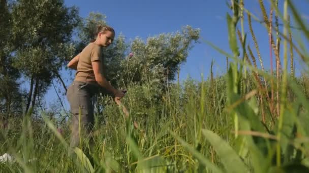 Jovem cortando a grama verde com a foice no campo de verão junto ao rio. Bela paisagem. Trabalha no terreno. Mulher rural real . — Vídeo de Stock