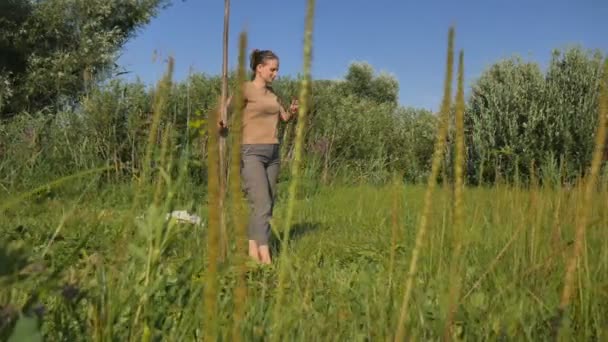 Mujer joven cortando la hierba verde con la guadaña en el campo de verano junto al río. Hermoso paisaje. Trabaja en el campo. Mujer rural real . — Vídeos de Stock