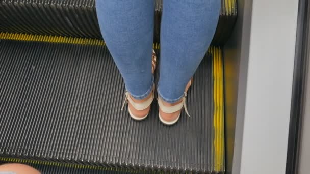 De benen van een vrouw in jeans, in zomerschoenen, sandalen op een bewegende roltrap. — Stockvideo