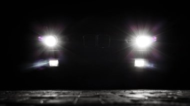 Geceleyin karanlığın içinden geçen klasik bir arabanın parlak farları. Asfaltta ışınlar ve mercekler parlıyor. Stilize gece çekimi.