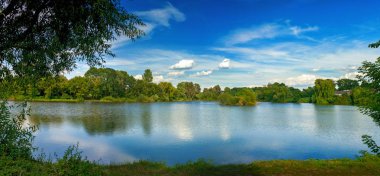 Bir gölde, canlı gökyüzü, beyaz bulutlar ve ağaçlar sakin manzara temiz mavi suya yansıyan
