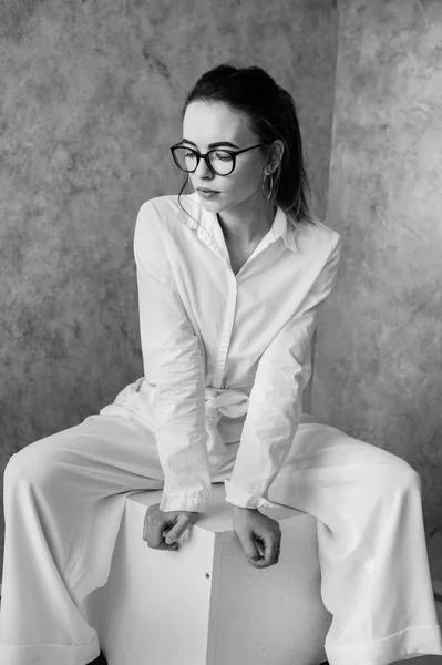 年轻女子戴着眼镜坐在工作室的白色立方体上，与灰蒙蒙的背景隔离开来，从相机上看. — 图库照片
