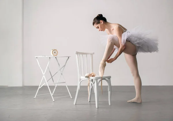 Bailarina de balé em um vestido branco com cadeira amarrando seu deslizamento de balé — Fotografia de Stock