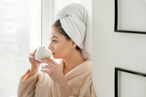 Chica joven con una toalla en la cabeza está bebiendo con taza de café, cerca del alféizar de la ventana en el baño —  Fotos de Stock