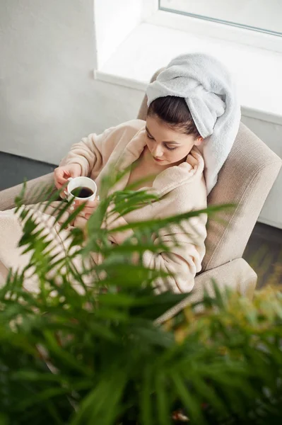 Chica en un albornoz y toalla en la cabeza se sienta relajarse en una silla con café —  Fotos de Stock