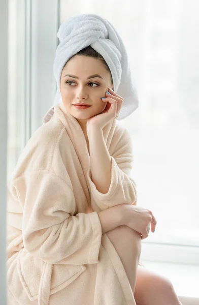 Porträtt av vacker flicka och handduk på huvudet nära fönstret, hem stil avslappnings koncept efter dusch. — Stockfoto