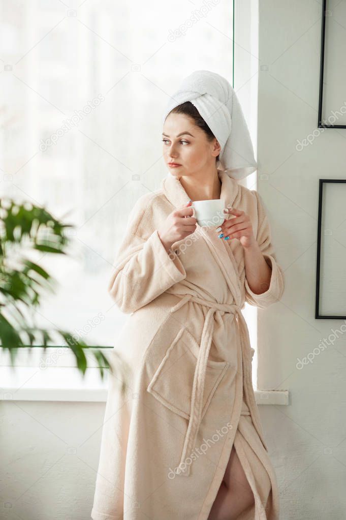 Attractive young girl in bathrobe and with a towel on her head is holding a cup, sitting on a window sill in the bathroom