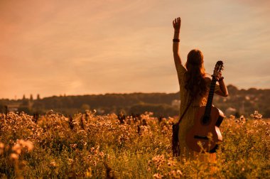 Gün batımının sıcacık ışığında bir tarlada gitar tutan bohem tarzı bir kadın silüeti.
