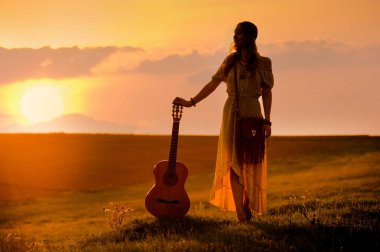 Siluet 'te bohem tarzı giyinen bir kadın gitar tutuyor.