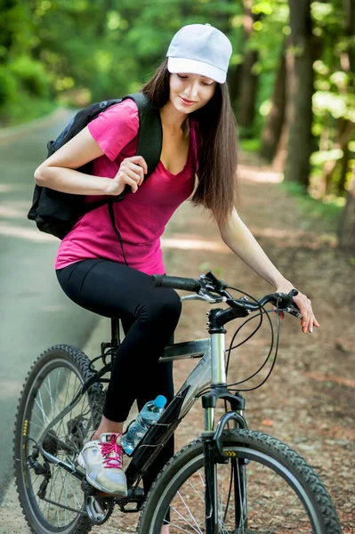 在森林里的沥青路面上骑自行车的女骑手 戴着白帽 — 图库照片