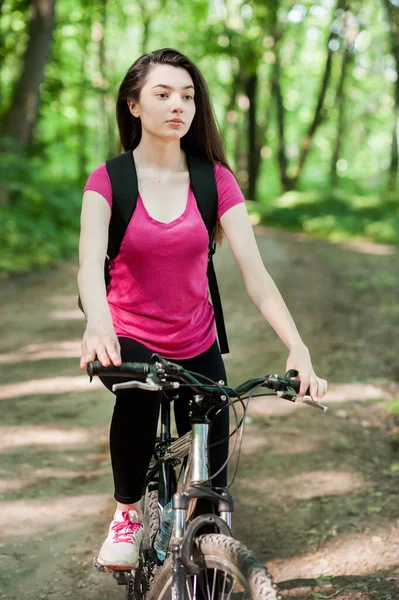 在森林里的沥青路面上骑自行车的女骑手 穿着粉色T恤 — 图库照片