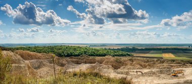 Landscape with quarry and excavator working and fields under blue sky with clouds.