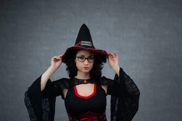 Siyah ve kırmızı kostümlü, iki eli başının üzerinde cadı şapkası tutan çekici bir cadı. Cadılar Bayramı için ideal, şeker mi şaka mı, fantezi bayramı. Gri arkaplanda kopya alanı olan yatay stüdyo portresi.