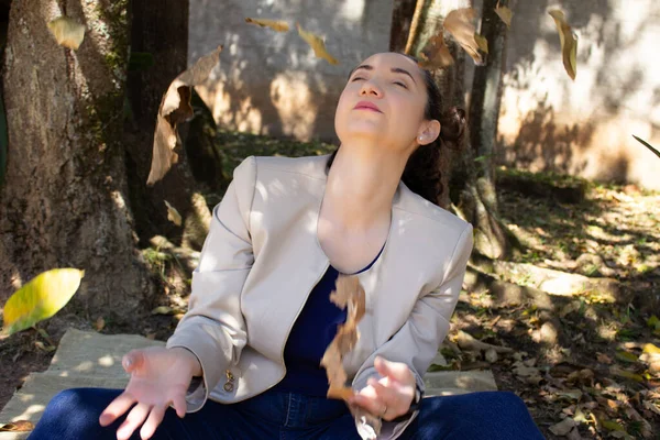 Parkta yerde oturan bir kadın, düşen yaprakları yakalıyor. Sonbahar atmosferi, doğal yaşam ve çevresel kaygılar için idealdir. Açık havada, güneş ışığıyla yatay portre.