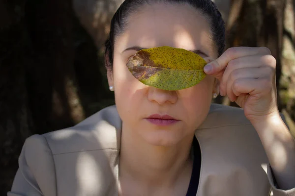 İki gözünü de solmuş ve lekeli bir yaprakla gizleyen bir kadın. Çevresel farkındalık, sürdürülebilirlik, botanik, sonbahar, sonbahar sezonu için ideal. Fotokopi alanı ile açık havada yatay portre.