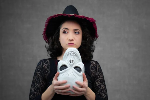 Şüpheli cadı baş aşağı bir kafatası tutuyor, tek kaşını kaldırmış başka tarafa bakıyor. Cadılar Bayramı için ideal, Ölüler Günü, ürkütücü temalar ve peri masalları için kötü adam. Kopya alanı ile yatay stüdyo portresi.