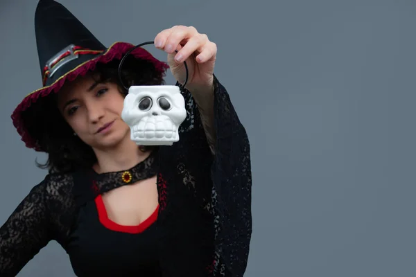 Yakışıklı dost canlısı cadı tek eliyle plastik bir kafatası sepeti tutuyor. Cadılar Bayramı için ideal, şeker mi şaka mı, cadılık, büyü ve fantezi teması. Gri arkaplanda kopyalama alanı olan yatay portre.