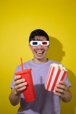 Üç boyutlu gözlüklü genç adam, bir kutu patlamış mısır ve sarı arka planda kırmızı soda bardağını gösterirken gülümsüyor. Fotokopi alanı, sinema, eğlence ve eğlence konsepti.