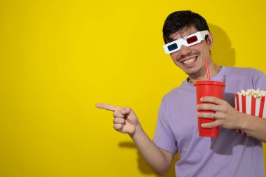 Üç boyutlu gözlüklü gülümseyen adam elinde bir kutu patlamış mısır ve sarı arka planda kırmızı soda bardağı tutarken, sinema ve eğlence kavramını işaret ediyor.