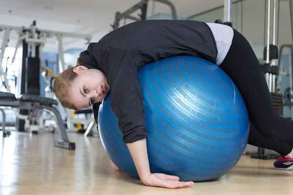 Spor salonunda yorgun, yorgun, sıkılmış ve yorgun görünümlü mavi jimnastik topu üzerinde yatan genç beyaz kadın