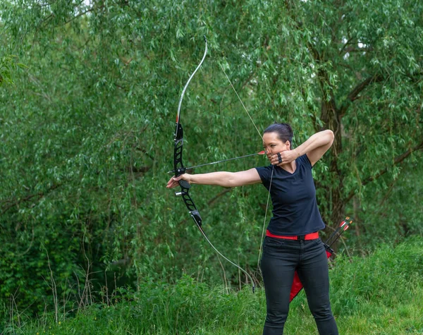 Tiro con arco en la naturaleza. Una joven y atractiva mujer está entrenando en un tiro de arco con una flecha en un objetivo en el bosque —  Fotos de Stock