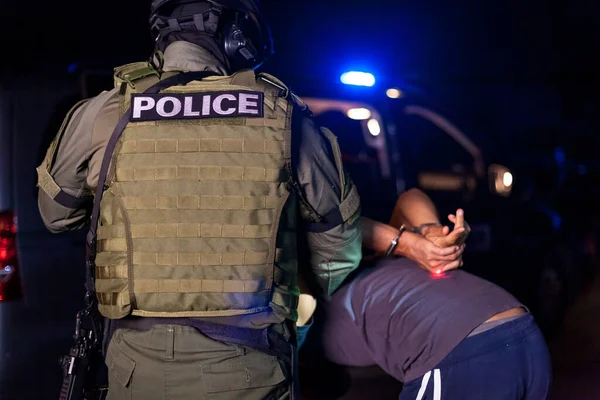 Un oficial de policía pone esposas en las manos de un delincuente durante un arresto. Coche de policía con faros intermitentes. Borrosa. — Foto de Stock
