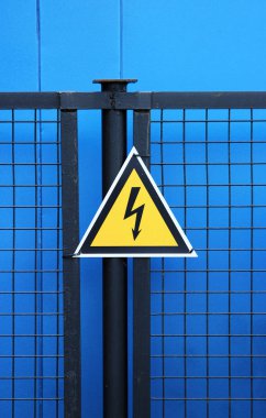 Yüksek gerilim üçgen uyarı işareti mavi bir arka plan üzerinde bir metal kafes çit üzerine monte