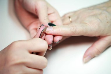 Tırnak bakımı manikürcüsü yaşlı bir kadına metal bir alet mızrağıyla manikür yapıyor. Çivinin yüzeyini düzleştiriyor. Ateş edildiğini bildiriyor.