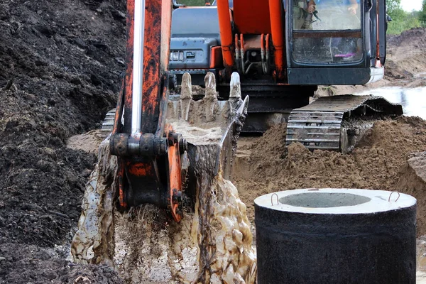 The excavator draws water from the ditch with a bucket where the road will pass. rain season. repair and expansion of the road. storm concrete rings. Gatchina, Russia, reportage. — Stock Photo, Image