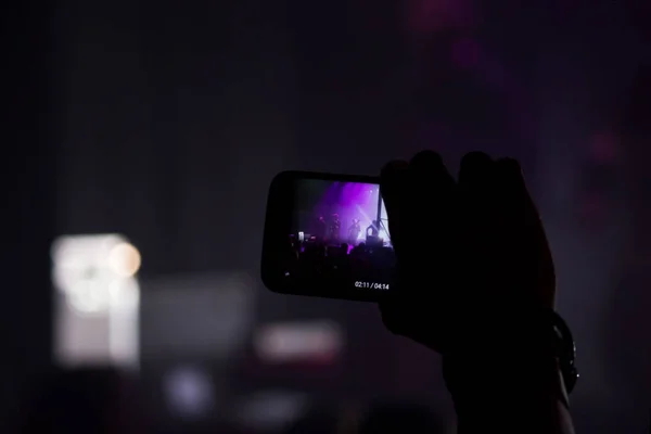 Man hand with a smartphone records a live concert of the group consisting of four cellists and a drummer. The scene is lit with purple spotlights — Stock Photo, Image