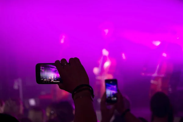 Mão de homem com um smartphone grava um concerto ao vivo do grupo composto por quatro violoncelistas e um baterista. A cena é iluminada com holofotes roxos — Fotografia de Stock