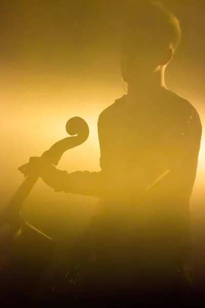 Musicista maschile con violoncello sul palco illuminato da una luce giallo miele da dietro. reportage — Foto Stock