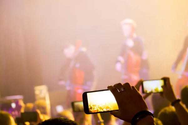 Mão de homem com um smartphone grava um concerto ao vivo do grupo composto por quatro violoncelistas e um baterista. A cena é iluminada com holofotes roxos — Fotografia de Stock