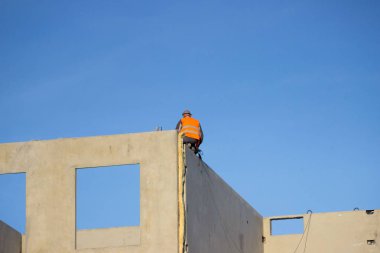 Profesyonel inşaat işçisi inşaat halindeki bir evin duvarının kenarında oturuyor ve vinç tarafından sanayi bölgesinde taşınan levhayı bekliyor. Beşinci kat. güvenlik ihlali