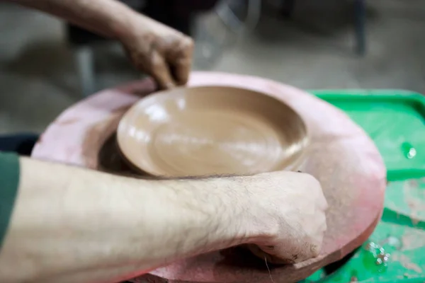 A master ceramist with 13 years of experience made plate from red clay on a potter's wheel, on a sheet of particle board for better removal and further drying. before removed chipboard he cuts plate o — Stock Photo, Image