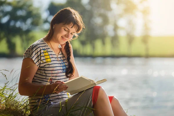 beautiful woman reading a book in the nature.brunette in a striped T-shirt reading a book against the background of a river