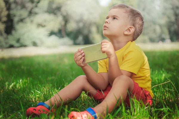 A little boy is sitting with the phone on the grass.little boy in a yellow T-shirt and red shorts sitting on the grass with a smartphone