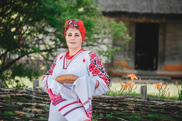 a Ukrainian woman in traditional attire holds rushnyk with bread and salt on the background of an old house and flowers.a woman greets guests according to tradition