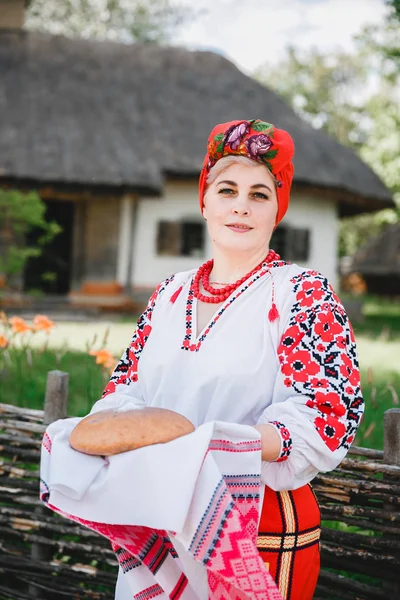 一位穿着传统服装的乌克兰妇女在老房子和鲜花的背景上 Rushnyk 面包和盐 一个女人根据传统问候客人 — 图库照片