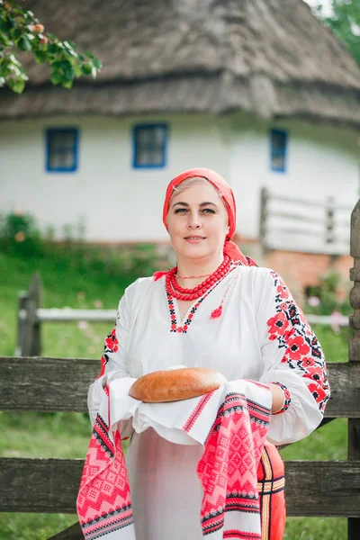 一位穿着传统服装的乌克兰妇女在老房子和鲜花的背景上 Rushnyk 面包和盐 一个女人根据传统问候客人 — 图库照片