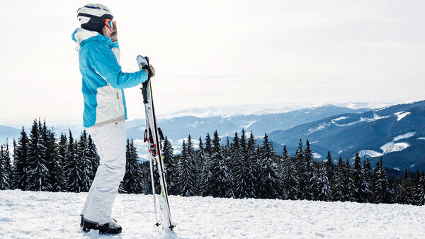 a woman in a blue suit, a helmet and glasses is standing with skis on top of a mountain.
