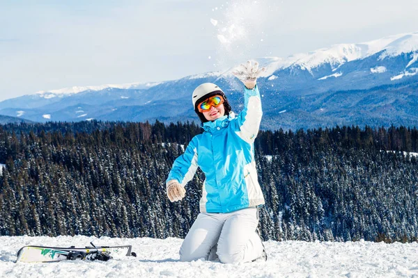 穿着蓝色西装 头盔和眼镜的妇女坐在山顶上滑雪板旁的雪地上 女孩把雪扔在相机上 — 图库照片