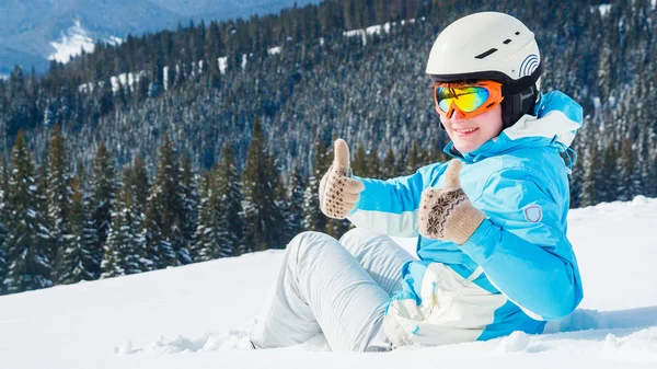 穿着蓝色西装 头盔和眼镜的妇女坐在山顶上滑雪板附近的雪地上 — 图库照片