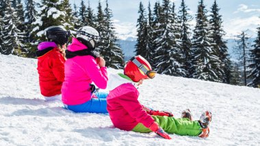 Bir grup insan kırmızı Kayak takım elbiseli bir dağın tepesinde kar üzerinde oturuyor. Bir aile kayakçı kar üzerinde oturmuş dinleniyor